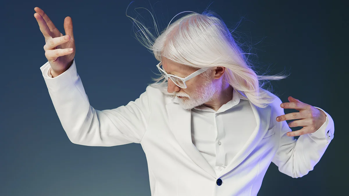 A middle aged albino man with long white hair in a white suit, looking faulous and dancing